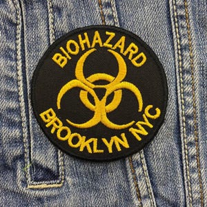 Könnte beinhalten: Schwarzer und gelber gestickter Aufnäher mit dem Text "BIOHAZARD" und "BROOKLYN NYC" um ein Biohazard-Symbol.