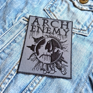 Könnte beinhalten: Ein grauer rechteckiger Aufnäher mit den Worten "ARCH ENEMY" in fetten, schwarzen Buchstaben. Der Aufnäher zeigt eine schwarze Grafik eines Schädels und andere Designelemente. Der Aufnäher ist auf eine blaue Jeansjacke genäht.