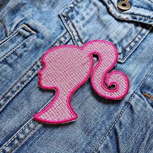 Può includere: Toppa ricamata rosa da stirare con la silhouette di una donna con una coda di cavallo. La toppa è su una giacca di jeans blu.
