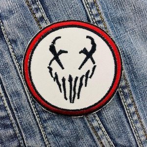 May include: A white patch with a black skull design and a red border. The skull has two eyes and a mouth with teeth. The patch is on a blue denim background.