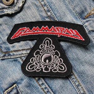Pode incluir: Um remendo bordado preto e vermelho com o texto "GAMMARAY" em um arco vermelho sobre um símbolo triangular preto e branco.