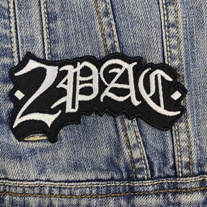 Puede incluir: Un parche bordado en blanco y negro con la palabra "2PAC" en un estilo de fuente gótica. El parche está cosido en una chaqueta vaquera azul.