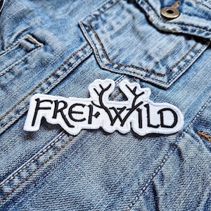 Puede incluir: Parche blanco bordado con letras negras que dice "FREY WILD" con dos astas estilizadas sobre el texto. El parche está cosido en una chaqueta vaquera azul.