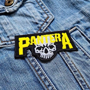 May include: A black and yellow embroidered patch with the word "PANTERA" and a skull design. The patch is rectangular and sewn onto a denim jacket.