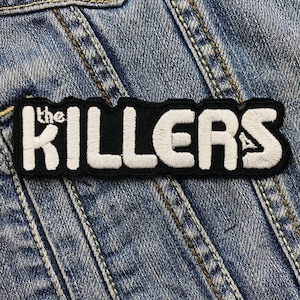 May include: Embroidered patch featuring the words "the KILLERS" in white on a black background. The patch is rectangular and is sewn onto a blue denim jacket.