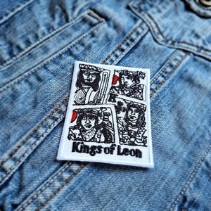May include: A white embroidered patch featuring four stylized portraits with red accents, and the text "Kings of Leon" in black. The patch is placed on a denim jacket.