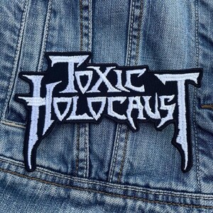 Może przedstawiać: Czarna i biała naszywka z napisem "TOXIC HOLOCAUST" w stylizowanej czcionce. Naszywka jest prostokątna z postrzępioną krawędzią i jest umieszczona na dżinsowej kurtce.
