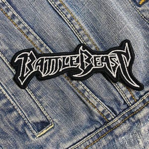 May include: A black and white embroidered patch featuring the words "BATTLE BEAST" in a decorative font. The patch is on a blue denim background, with visible stitching and a rectangular shape.