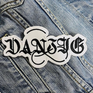 Op de afbeelding: Een wit geborduurd embleem met het woord "DANZIG" in zwarte gotische letters. Het embleem is afgebeeld op een spijkerjasje, wat het ontwerp en de mogelijke plaatsing op kleding benadrukt.