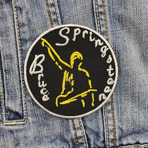 May include: Black and white embroidered patch with a yellow silhouette of a person playing guitar. The text "Bruce Springsteen" is written around the edge of the patch.