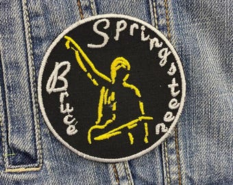 May include: Black and white embroidered patch with a yellow silhouette of a person playing guitar. The text "Bruce Springsteen" is written around the edge of the patch.