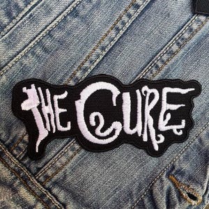 May include: Black and white embroidered patch with the text "The Cure" on a denim background.