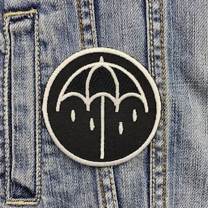 Könnte beinhalten: Schwarz-weiß gestickter Aufnäher eines Regenschirms mit Regentropfen auf blauem Jeansstoff.