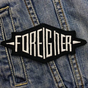 Op de afbeelding: Zwart-wit geborduurde patch met het woord "FOREIGNER" in een vet, gestileerd lettertype. De patch is genaaid op een blauw denim jack.