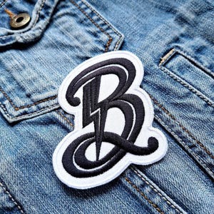 Peut inclure: Un patch brodé blanc et noir avec la lettre "B" dans une police stylisée. La lettre est soulignée en blanc et remplie de noir. Le patch est sur un fond en jean bleu.