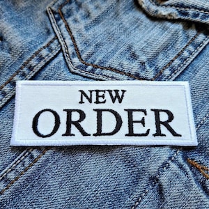 以下が含まれることがあります： 青いデニムの背景に「NEW ORDER」と黒文字で書かれた白い刺繍パッチ