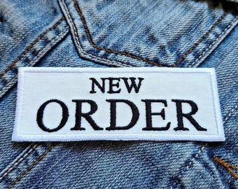 Può includere: Toppa ricamata bianca con testo nero che recita "NEW ORDER" su uno sfondo in denim blu.