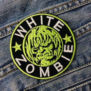 Peut inclure: Un écusson brodé noir et vert avec les mots "WHITE ZOMBIE" en lettres blanches et un visage de zombie vert au centre.