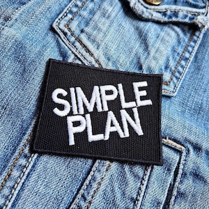 Può includere: Toppa ricamata in bianco e nero con il testo "SIMPLE PLAN" su una giacca di jeans.