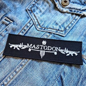 Pode incluir: Etiqueta bordada preta com costuras brancas apresentando o nome da banda "MASTODON" e um design estilizado.