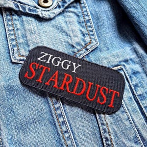 Pode incluir: Um patch retangular preto com as palavras "ZIGGY STARDUST" bordadas em branco e vermelho. O patch é costurado em uma jaqueta jeans azul clara, com costuras visíveis e um bolso.