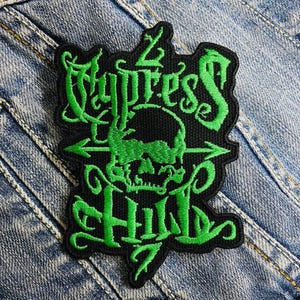 Puede incluir: Parche bordado verde con las palabras "Cypress Hill" y una calavera con huesos cruzados en el centro.