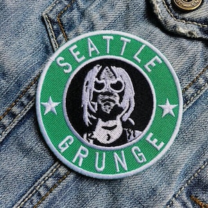 May include: A round, green and black patch with the words "SEATTLE GRUNGE" in white lettering. The patch features a stylized image of a figure with sunglasses and long hair. Two white stars are on either side of the figure.