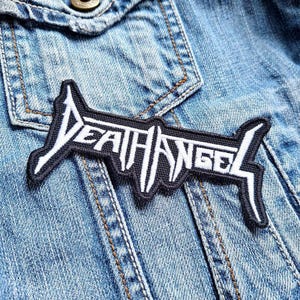 May include: A black and white embroidered patch with the text "DEATH ANGEL" in a stylized font. The patch is rectangular with a jagged border and is sewn onto a blue denim jacket.