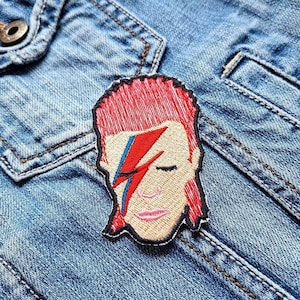 Könnte beinhalten: Gestickter Aufnäher mit einem stilisierten Porträt mit roten Haaren und einem roten und blauen Blitz auf dem Gesicht. Der Aufnäher ist auf einer hellblauen Jeansjacke angebracht, die das Design und die Farben zeigt.