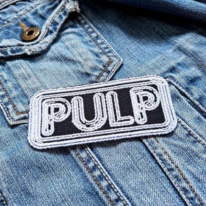May include: A black and white embroidered patch with the word "PULP" in white lettering on a black background. The patch is sewn onto a blue denim jacket.