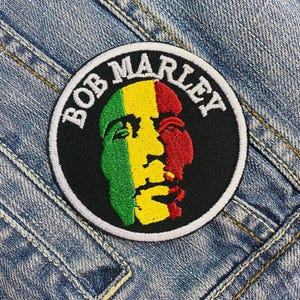 May include: A round patch featuring a portrait of Bob Marley, with the name "BOB MARLEY" in white lettering around the edge. The portrait is split vertically, with green, yellow, and red colors. The patch is set against a denim background.