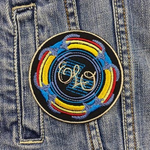 Puede incluir: Un parche bordado negro y azul con un círculo amarillo y rojo alrededor del exterior. El círculo tiene el texto "ELO" en una fuente cursiva blanca en el interior.