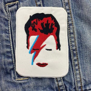 May include: White embroidered patch with a red and black image of David Bowie's face. The image is a close-up of his face with a lightning bolt across his eye.