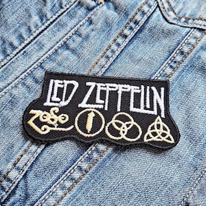 Könnte beinhalten: Schwarzer gestickter Patch mit dem Text "LED ZEPPELIN" in weißen Buchstaben. Der Patch zeigt außerdem vier goldene Symbole: ein stilisiertes "Zoso", einen Kreis mit einer Feder, einen Kreis mit einem dreiblättrigen Kleeblatt und ein Dreieck mit einem Kreis darin.