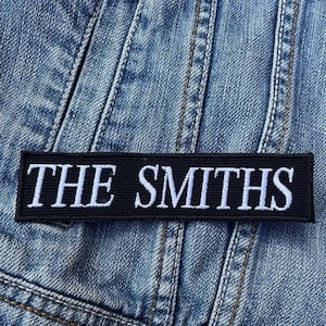 Puede incluir: Parche bordado en blanco y negro con el texto "THE SMITHS" sobre un fondo de mezclilla.