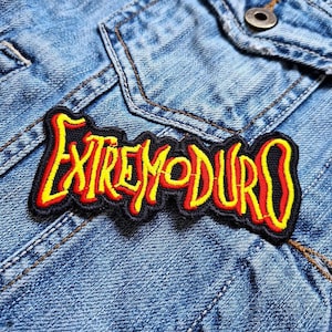 Puede incluir: Un parche bordado negro con el texto "EXTREMODURO" en amarillo y rojo, cosido sobre una chaqueta vaquera azul claro. El parche tiene un borde negro y está centrado en la zona del bolsillo de la chaqueta.