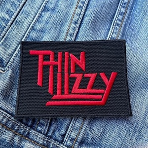 Può includere: Un cerotto in tessuto nero con il testo ricamato in rosso "Thin Lizzy" su uno sfondo in denim.