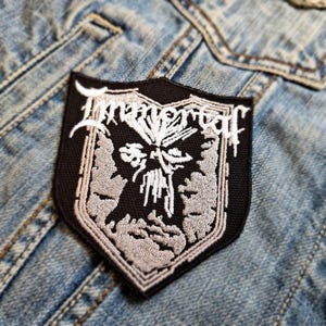 Puede incluir: Parche bordado en blanco y negro con la palabra "Immortal" en una fuente gótica. El parche presenta una imagen de calavera con un diseño detallado.