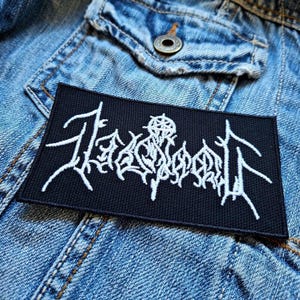 May include: Black embroidered patch with white lettering that reads "TÅRNSKOG" with a stylized symbol above the text.