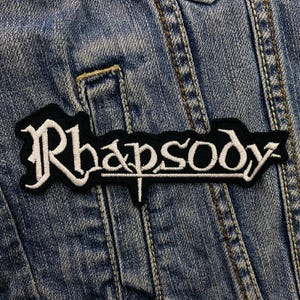 Op de afbeelding: Een geborduurde zwarte en witte patch met het woord "Rhapsody" in een gestileerd lettertype. De patch is genaaid op een blauw denim jack.