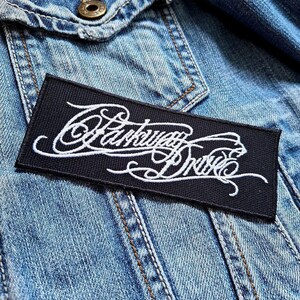 Może przedstawiać: Czarno-biała naszywka haftowana z tekstem "Parkway Drive" w ozdobnym, pochylonym kroju pisma. Naszywka jest przyszyta do niebieskiej dżinsowej kurtki.