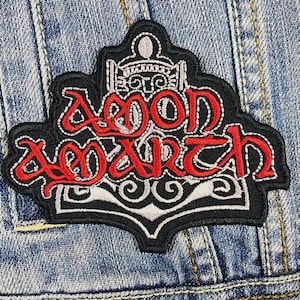 May include: A black and white embroidered patch with the words "Iron Maiden" in red lettering. The patch features a stylized design of a ship's anchor with a decorative border.