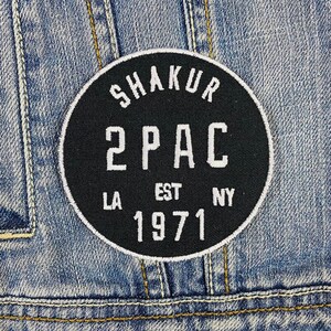Puede incluir: Parche bordado en blanco y negro con el texto "Shakur 2PAC EST 1971 LA NY" en un círculo.