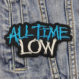 Könnte beinhalten: Ein gestickter schwarzer und weißer Aufnäher mit dem Text "ALL TIME LOW" in blauer Schrift. Der Aufnäher ist auf eine blaue Jeansjacke genäht.