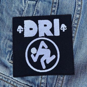 Op de afbeelding: Zwart-witte geborduurde patch met de tekst "DRI" en een gestileerd stickfiguur in een cirkel. De patch is op een blauwe denim achtergrond.