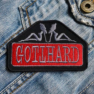 Op de afbeelding: Zwarte en rode geborduurde patch met het woord "GOTTHARD" in witte letters. De patch heeft een gestileerd ontwerp van twee figuren met hun armen boven hun hoofd.