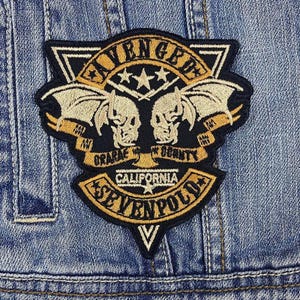 Może przedstawiać: Czarno-biała naszywka haftowana z napisami "Avenged Sevenfold" i "California" w stylu gotyckim. Naszywka zawiera również dwa skrzydła nietoperza z czaszkami oraz napisy "Orange County" i "Graffiti" na banerze.