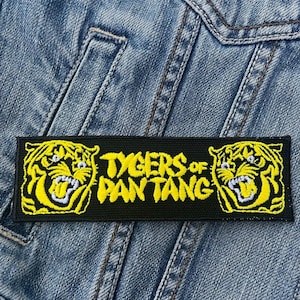 Op de afbeelding: Zwart rechthoekig embleem met gele tijgerkopafbeeldingen aan de uiteinden en de tekst "TYGERS of PANTANG" in het geel. Het embleem is op een spijkerjas genaaid.