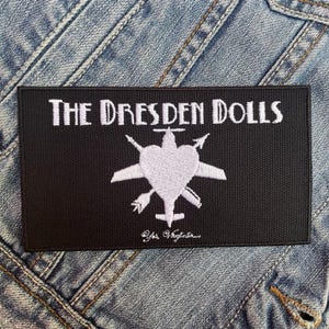 May include: Black embroidered patch with white text that reads "The Dresden Dolls" and a white silhouette of a plane with a heart in the middle. The text "Yes Virginia" is also embroidered in white below the plane.