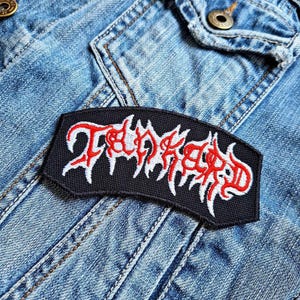 May include: A black, rectangular patch with the word "Tankard" embroidered in red and white, placed on a denim jacket. The text has a stylized, gothic font. The patch is a clothing accessory.